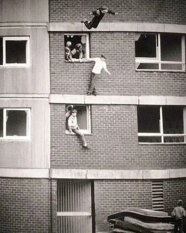 A black-and-white photo shows several children on the exterior of a multi-story brick building. One child is mid-air, having…