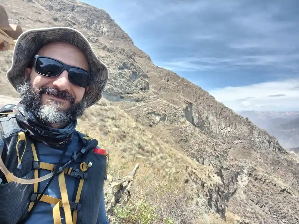 A man with a beard, sunglasses, and a bucket hat takes a selfie on a steep, rocky mountain slope. He wears a hydration pack…