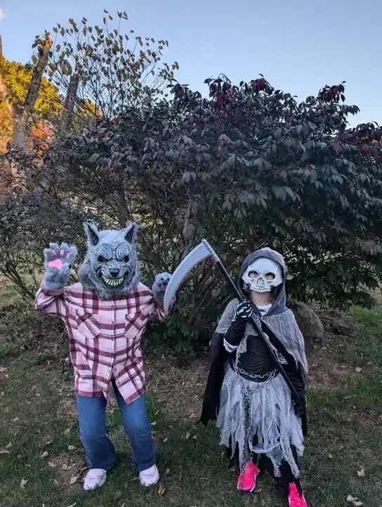 Two children in Halloween costumes stand outdoors in front of bushes. The child on the left wears a gray wolf mask with pink…