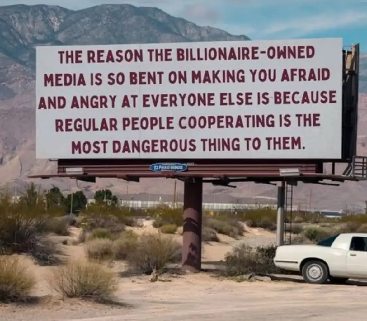 A large white billboard in a desert landscape with mountains in the background displays bold red text explaining that…