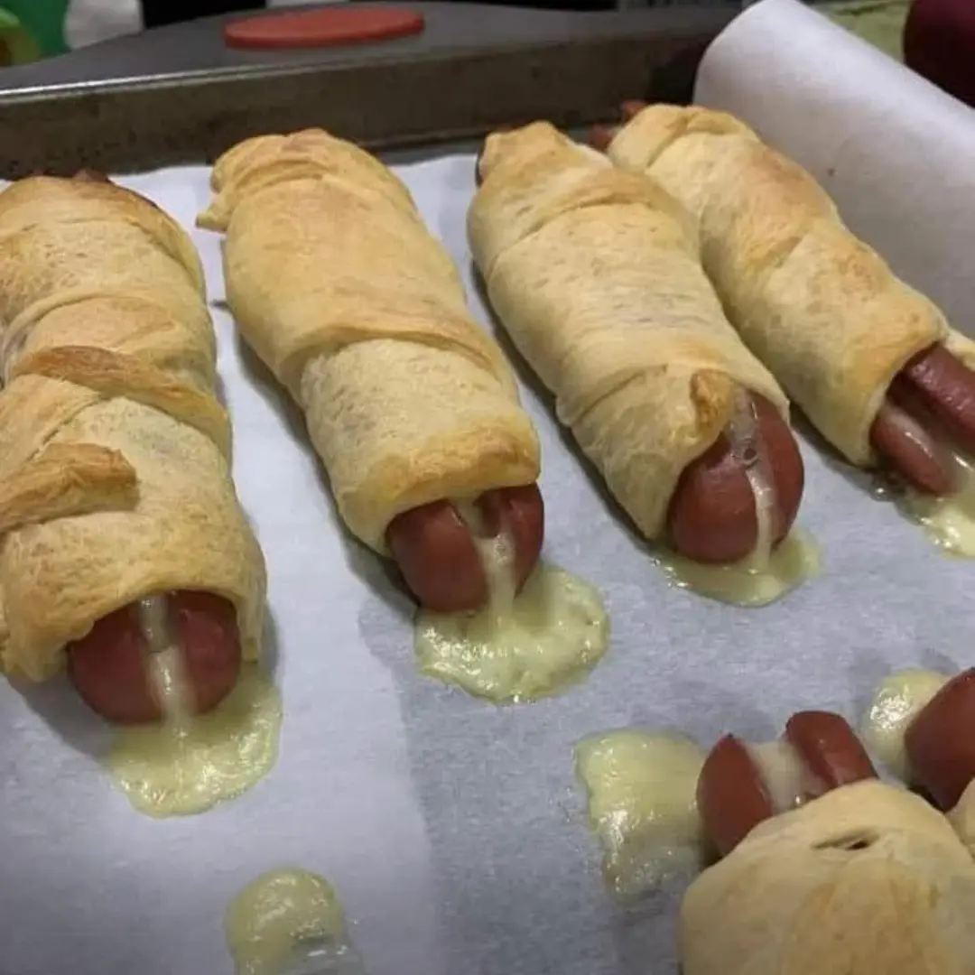 A close-up photo shows several baked hot dogs wrapped in golden-brown pastry, resting on parchment paper. Melted yellow…