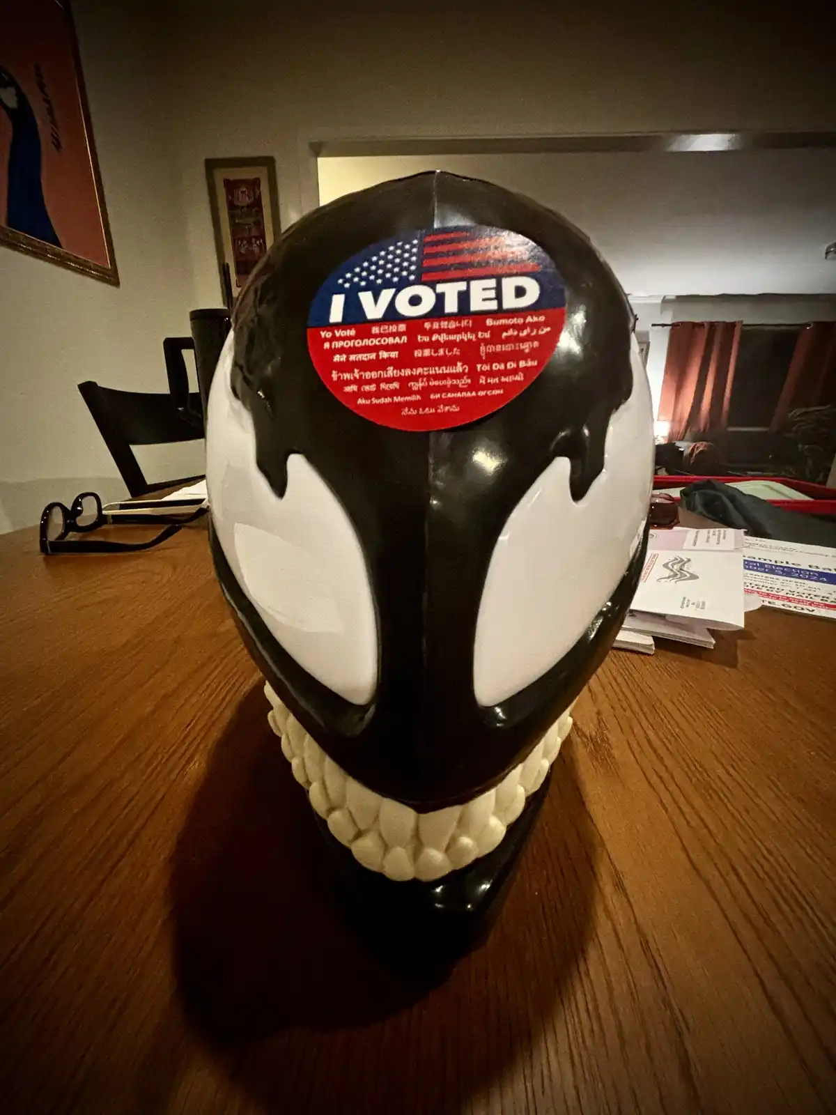 A black and white Venom mask sits on a wooden table with an 'I VOTED' sticker featuring the American flag and multilingual…