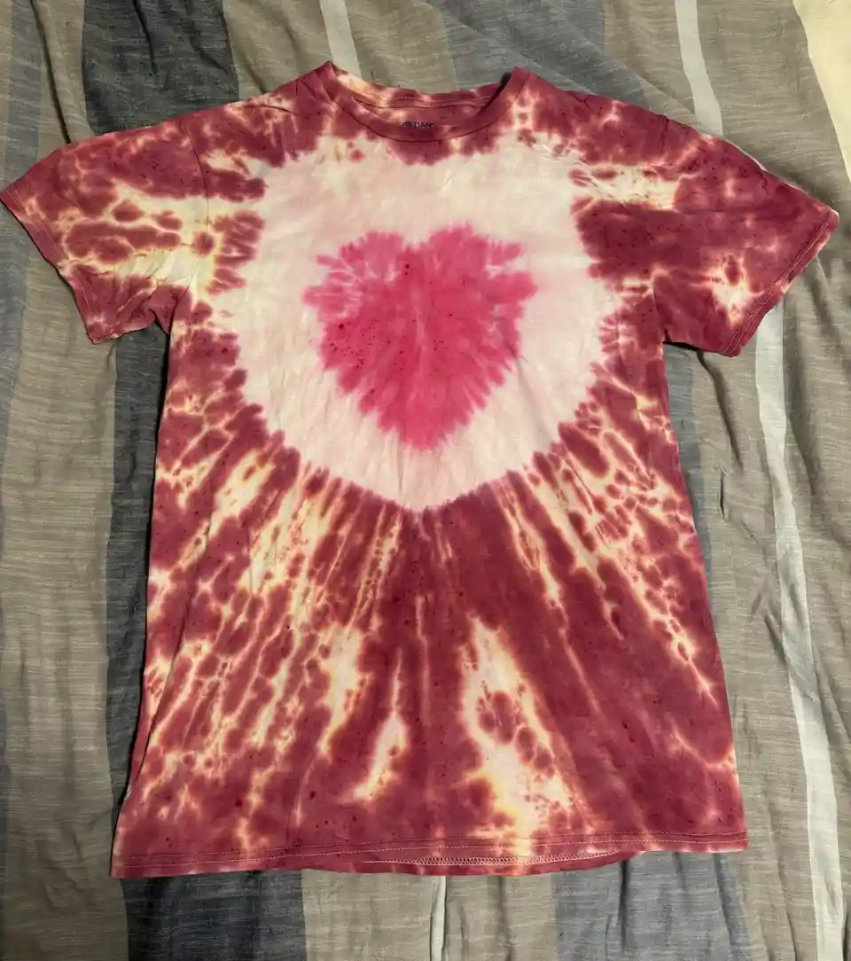 A red and white tie-dye t-shirt with a heart-shaped design in the center lies flat on a striped fabric surface. The shirt…