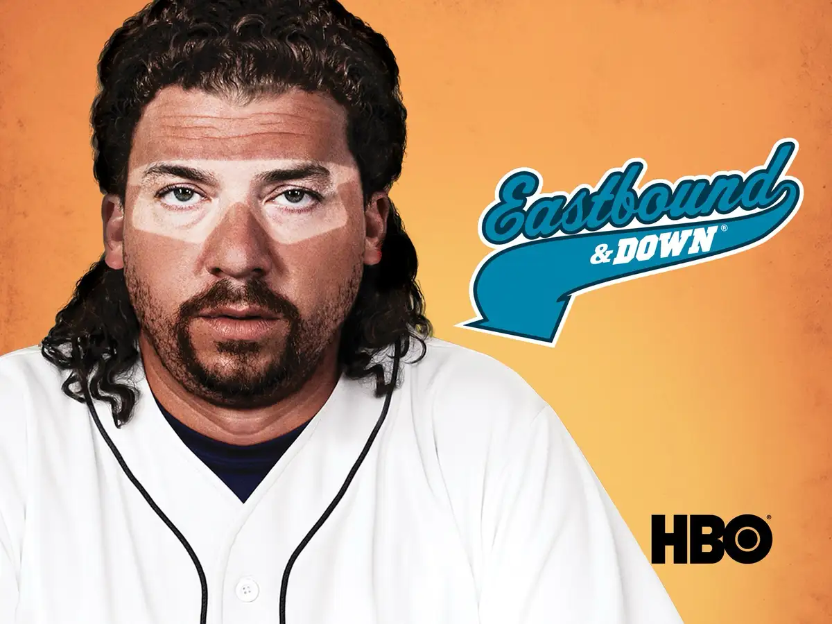 A man with long curly hair and a beard, wearing a white baseball jersey over a dark shirt, stares intensely at the camera…