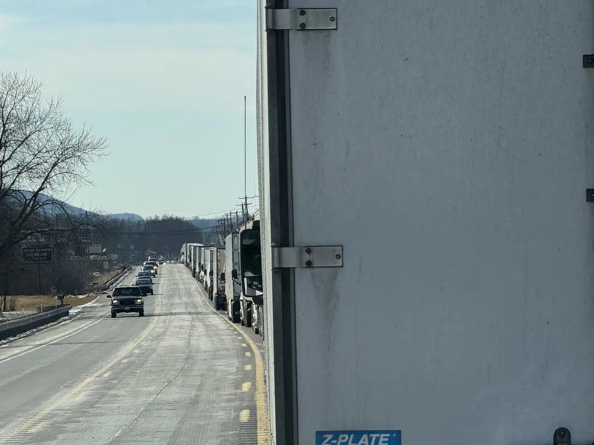 A photo taken from inside a truck shows a long line of semi-trucks parked along the right side of a road, with cars and…