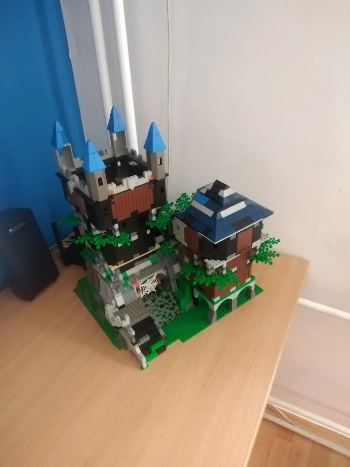 A detailed LEGO castle model with blue turrets, gray stone walls, and green foliage sits on a wooden table. The overlaid…