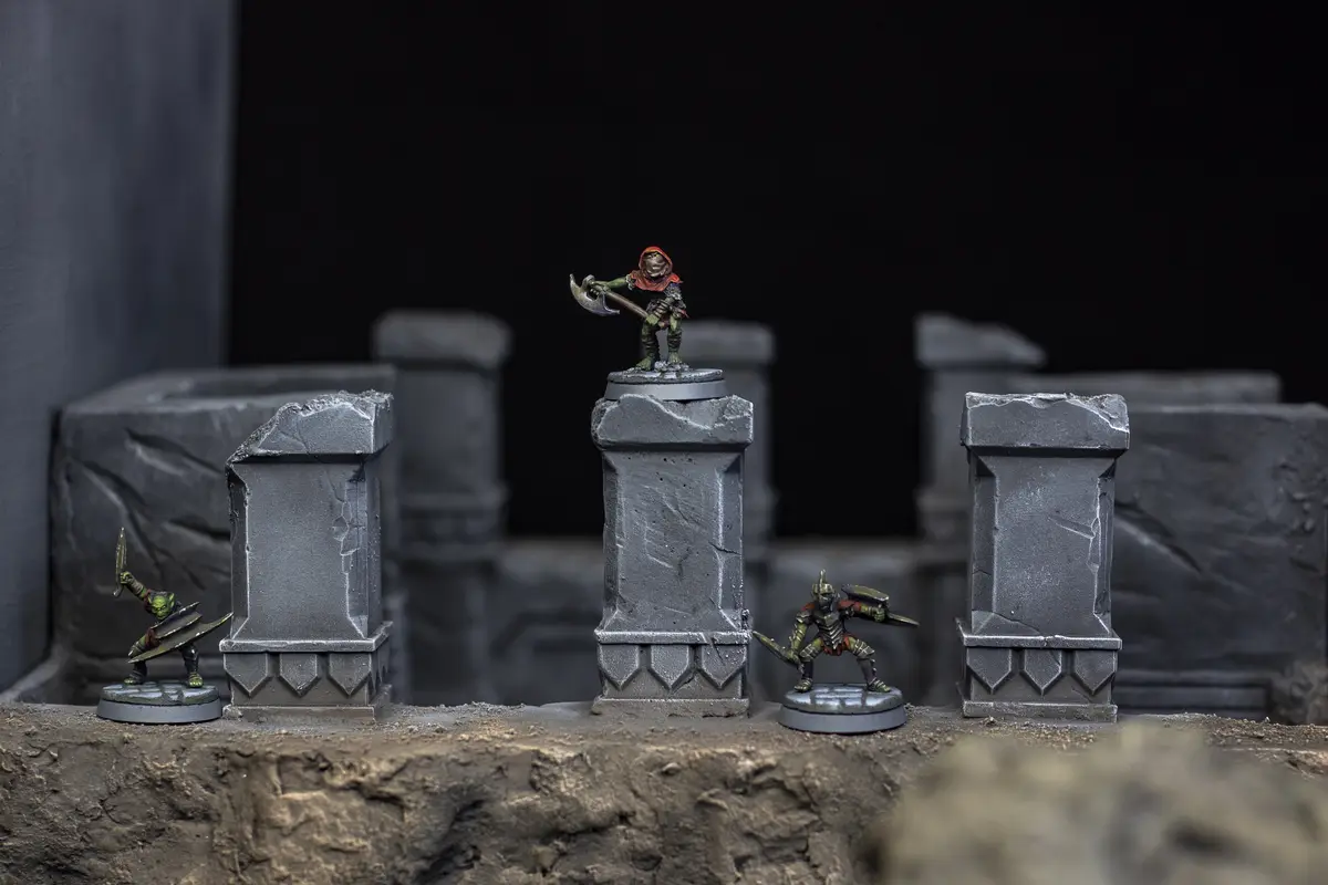 Three painted miniature fantasy figures, resembling goblins or orcs, are positioned on a diorama with stone pillars. One…
