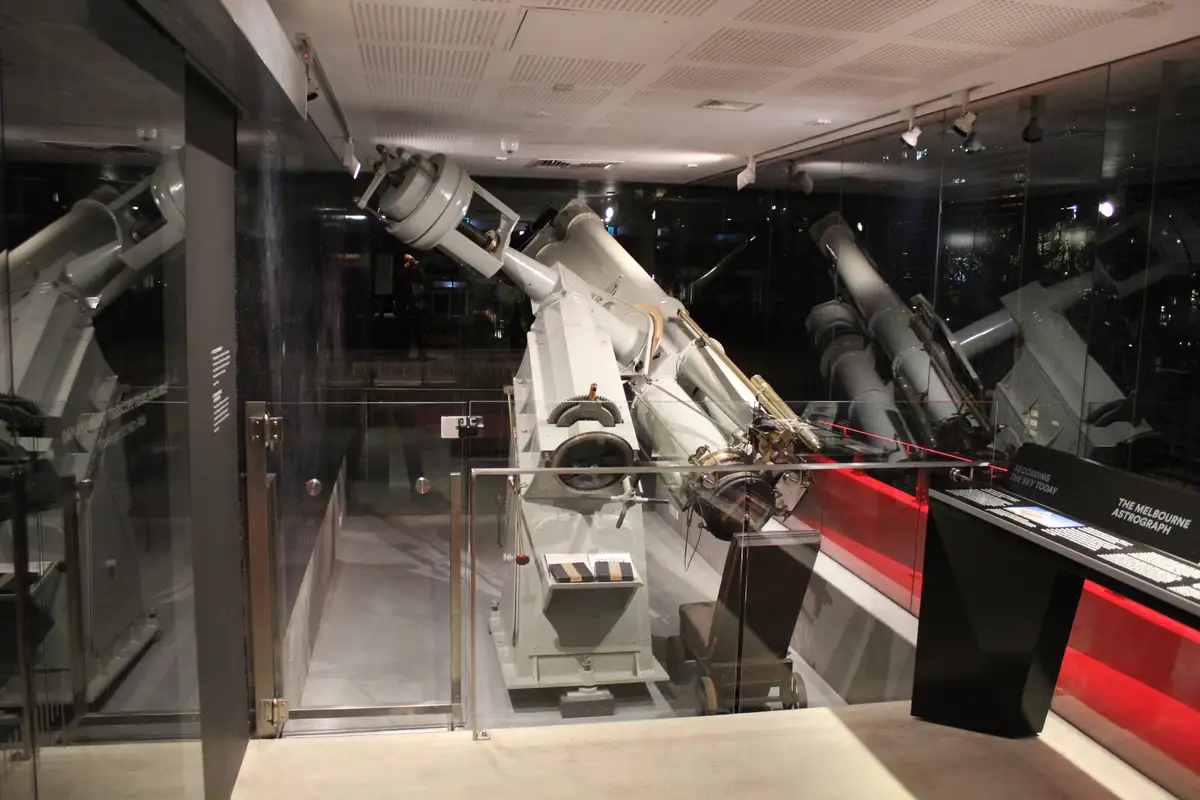 A large, white, vintage-style astronomical telescope, labeled 'THE MELBOURNE ASTROGRAPH', is displayed inside a…