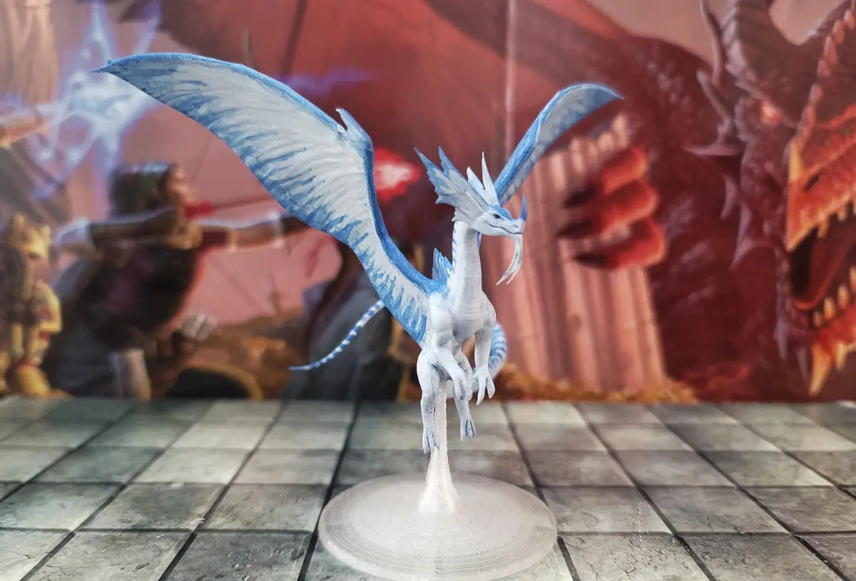 A small, blue-and-white painted dragon miniature stands on a clear plastic base on a checkered game board, with a blurred…