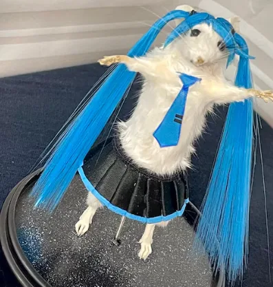 A white rat is dressed in a blue wig with twin tails, a blue tie, and a black pleated skirt, standing on a black circular…