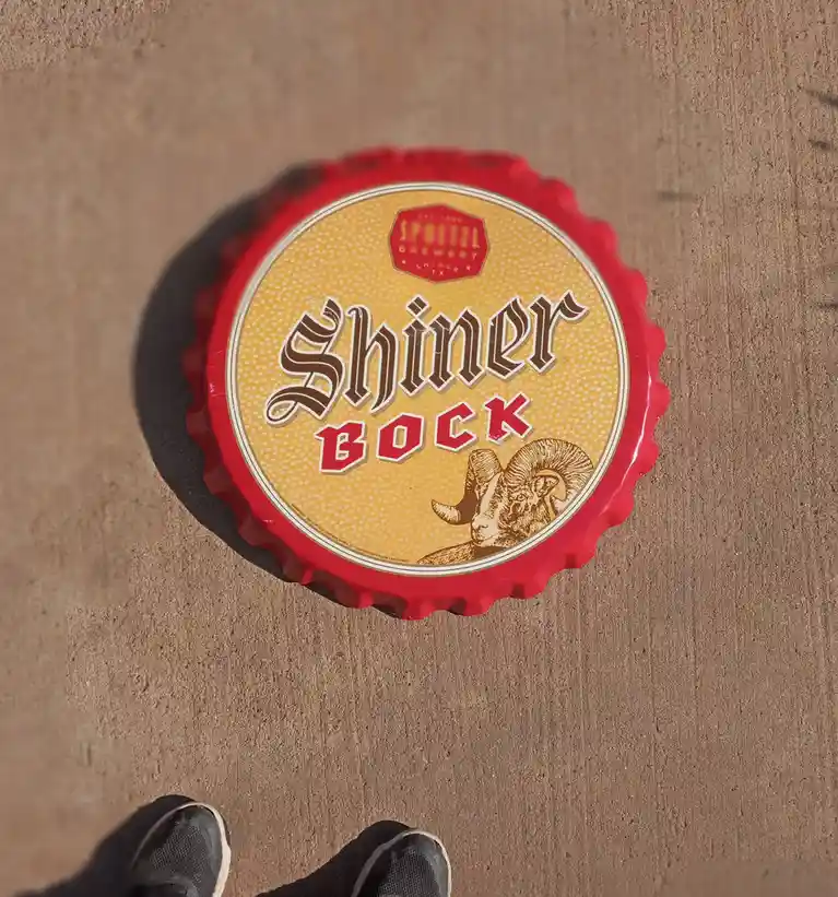 A red Shiner Bock beer bottle cap lies on a textured brown surface, with the brand name and ram logo visible. The bottom of…