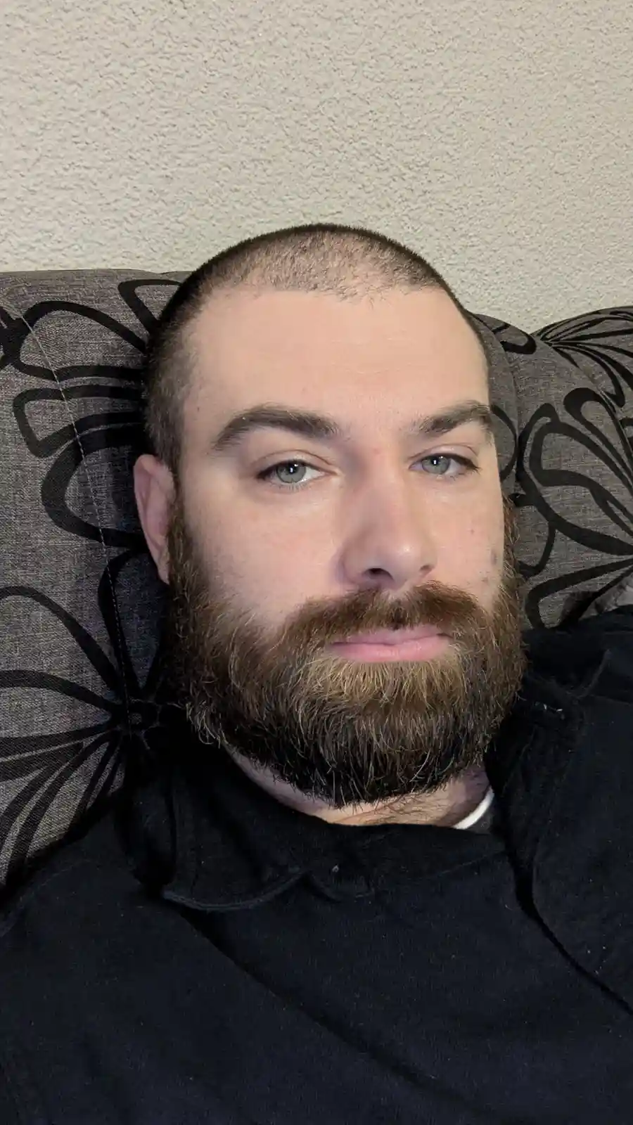 A man with a shaved head and full beard lies on a patterned couch, looking directly at the camera with a neutral expression…