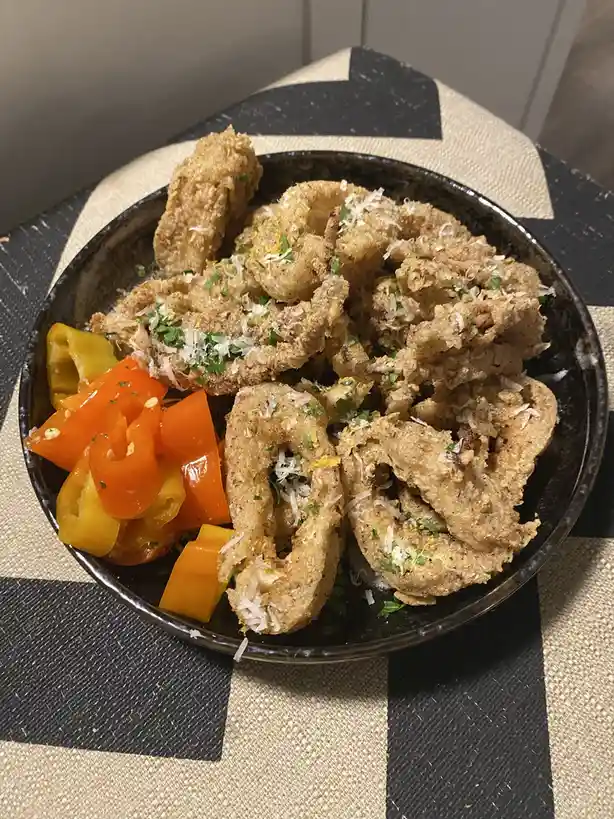 A dark bowl filled with fried calamari rings and chopped bell peppers sits on a black-and-beige checkered placemat. The…