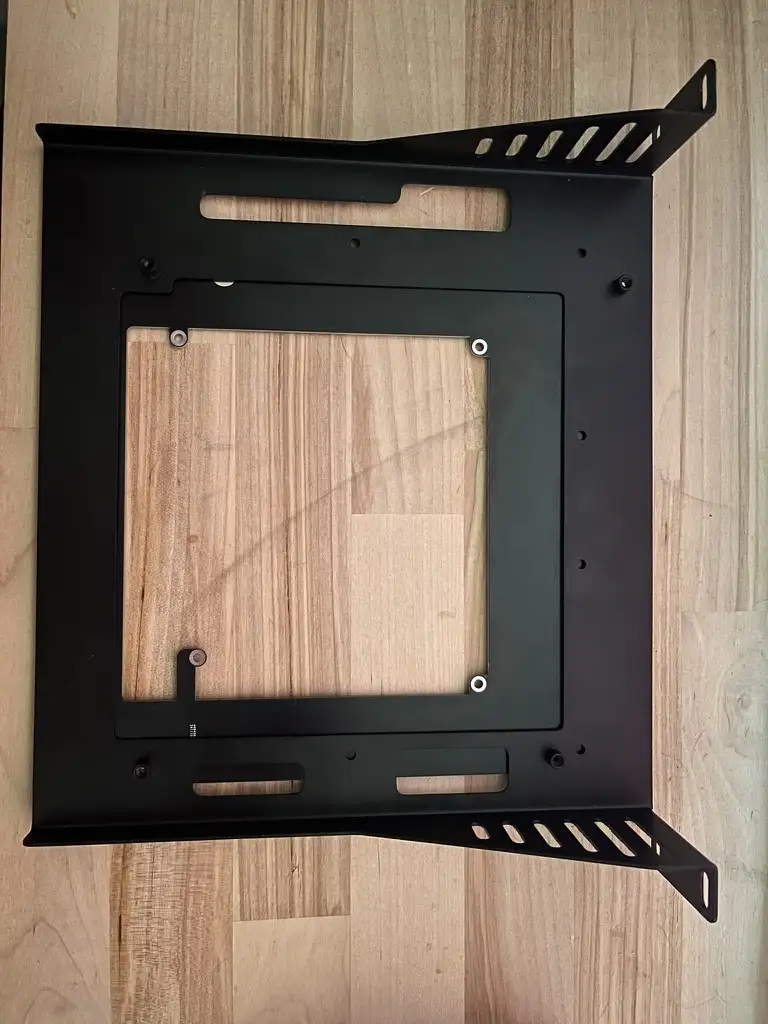A black metal TV wall mount frame is shown from above, sitting on a light wood floor. The mount is empty, revealing the wood…