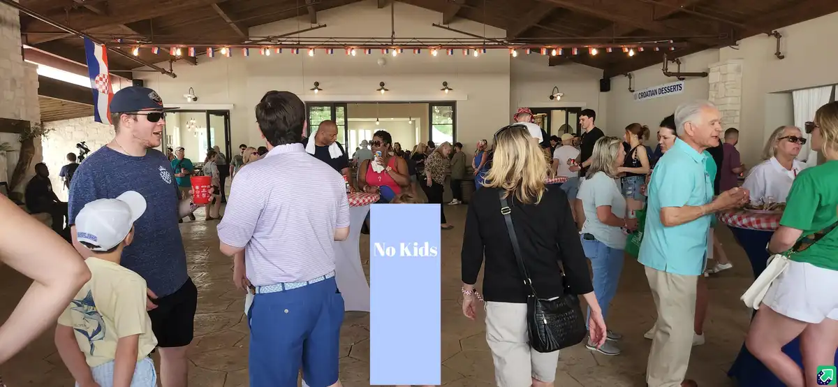 A crowded social gathering with adults mingling and eating at tables, but a young boy in a white cap and yellow shirt is…