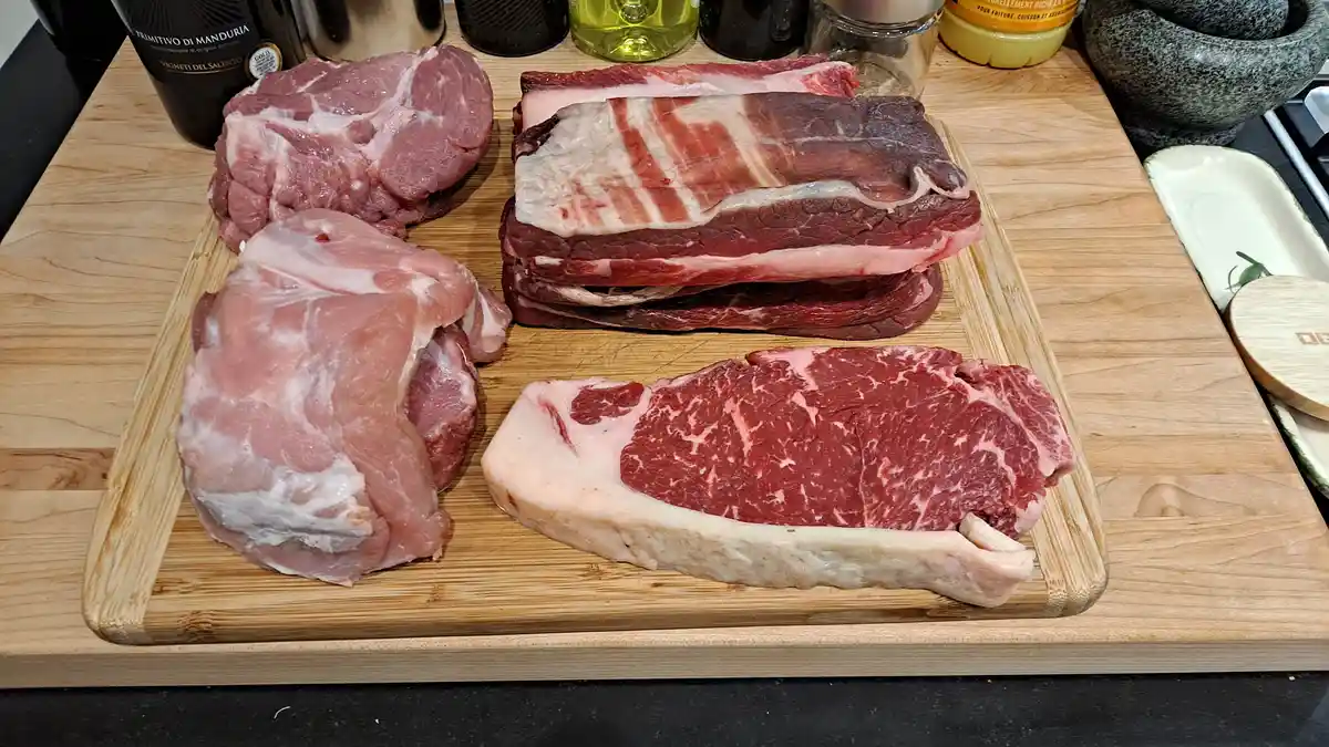 A wooden cutting board displays four raw meat cuts: two large pork pieces, a stack of beef ribs, and a thick beef steak with…