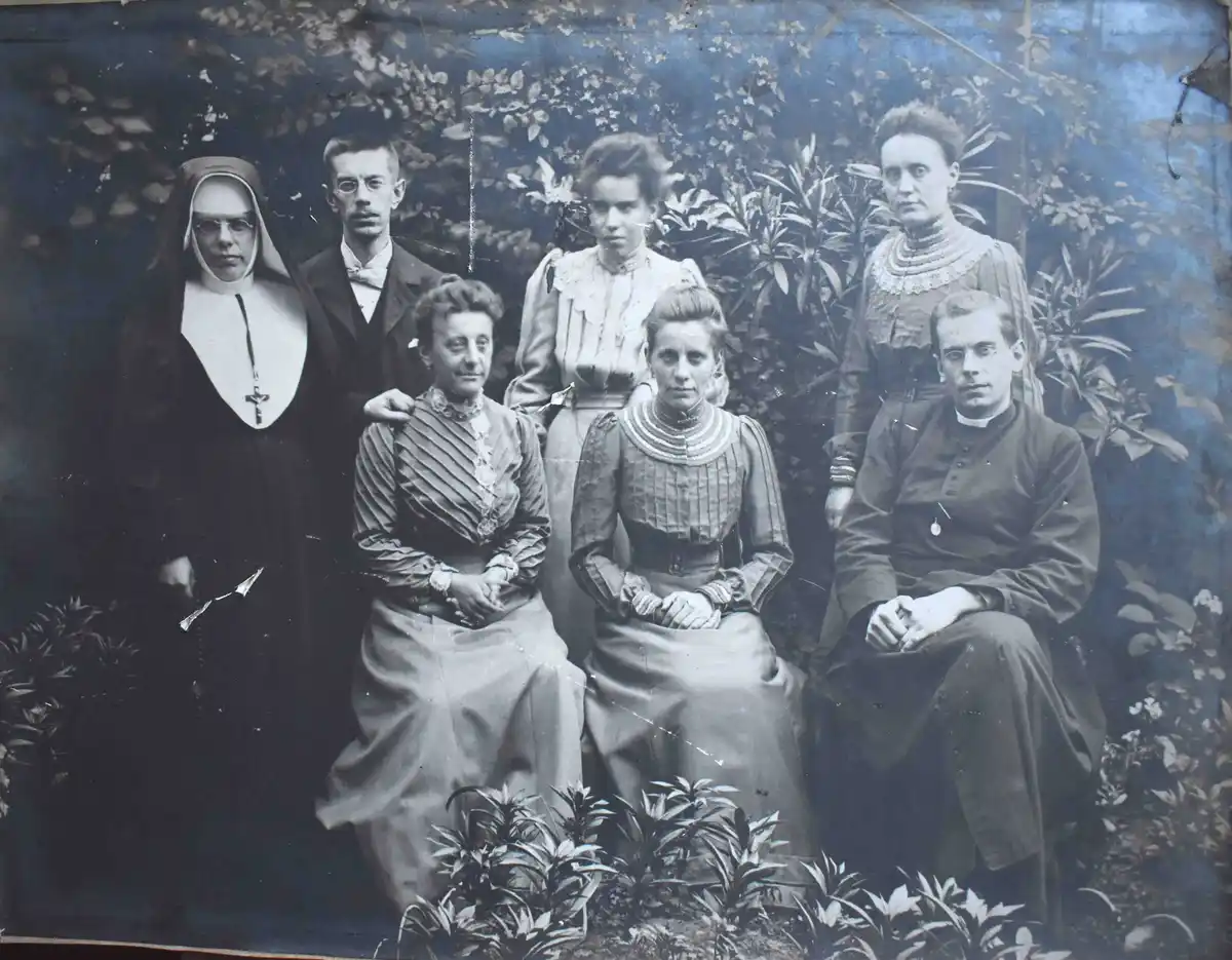 A vintage black-and-white photograph shows seven formally dressed people, including a nun, a priest, and several women in…