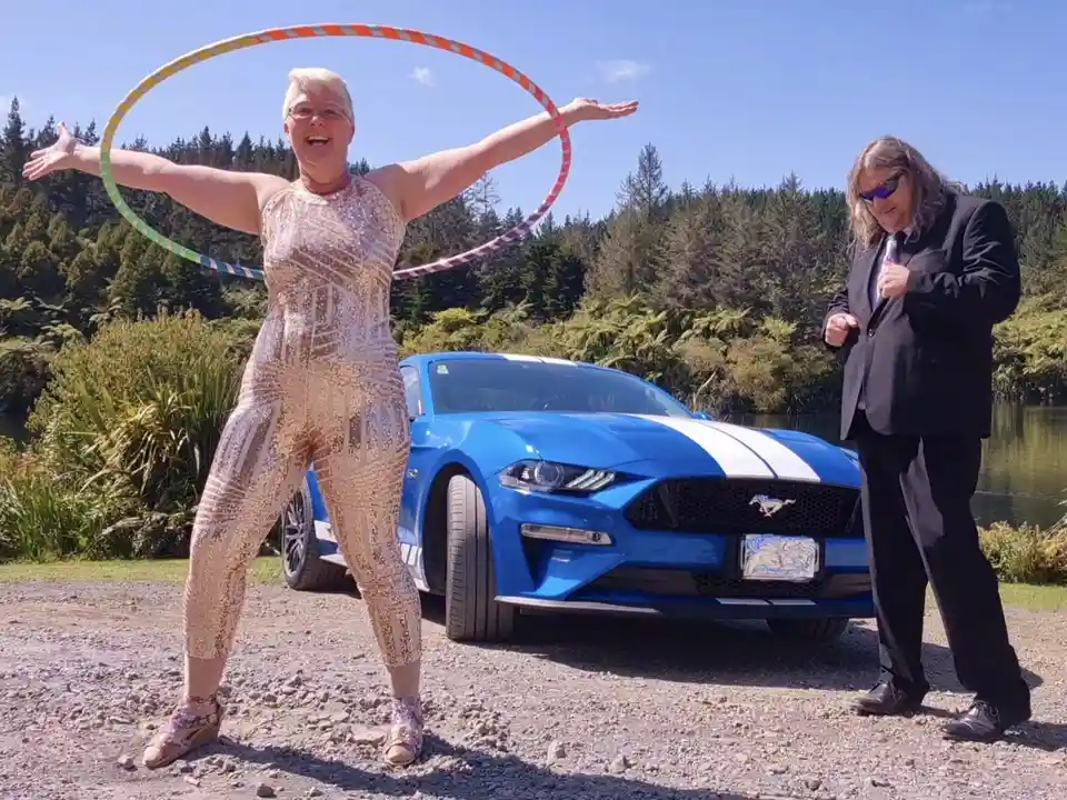 A woman in a sparkly gold jumpsuit joyfully spins a colorful hula hoop around her waist while standing in front of a blue…