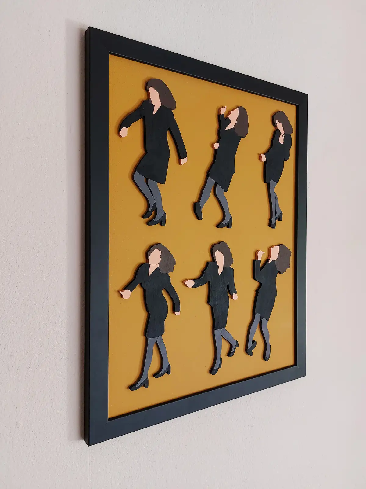 A framed piece of art on a wall shows six identical cutout figures of a woman in a black dress and heels, arranged in two…