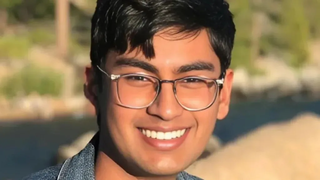 A young man with dark hair and glasses smiles broadly at the camera while wearing a denim shirt, with a blurred outdoor…