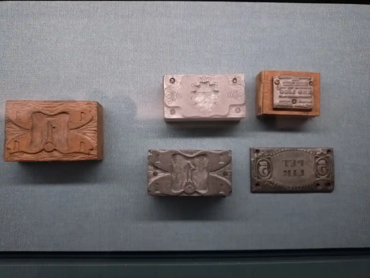 Five historical printing blocks for currency are displayed in a museum case on a blue fabric background. The blocks are made…