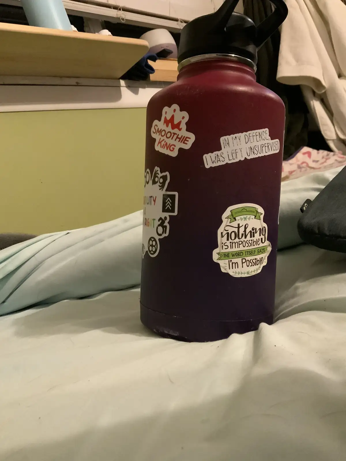A maroon water bottle sits on a bed, covered in various stickers including 'Smoothie King', 'Nothing is impossible', and the…