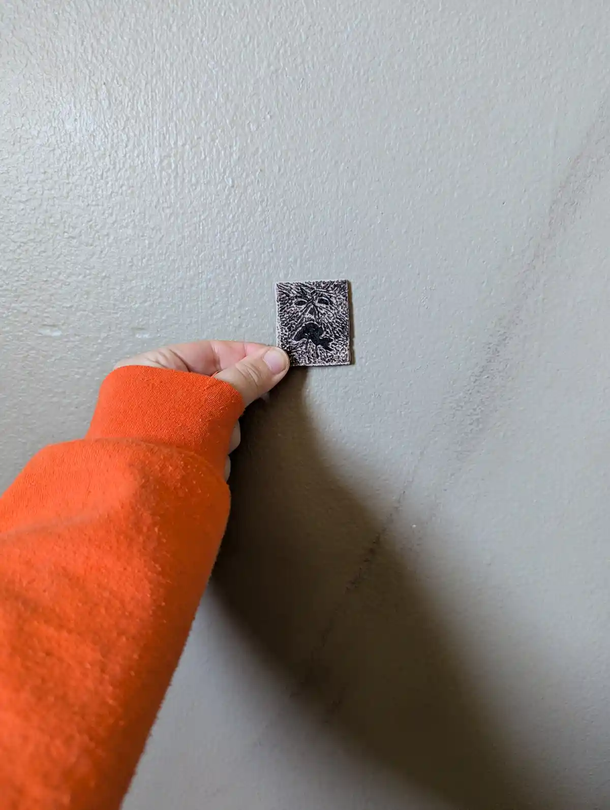 A hand in an orange sleeve holds a small square patch against a textured beige wall, revealing a faint, etched face on the…