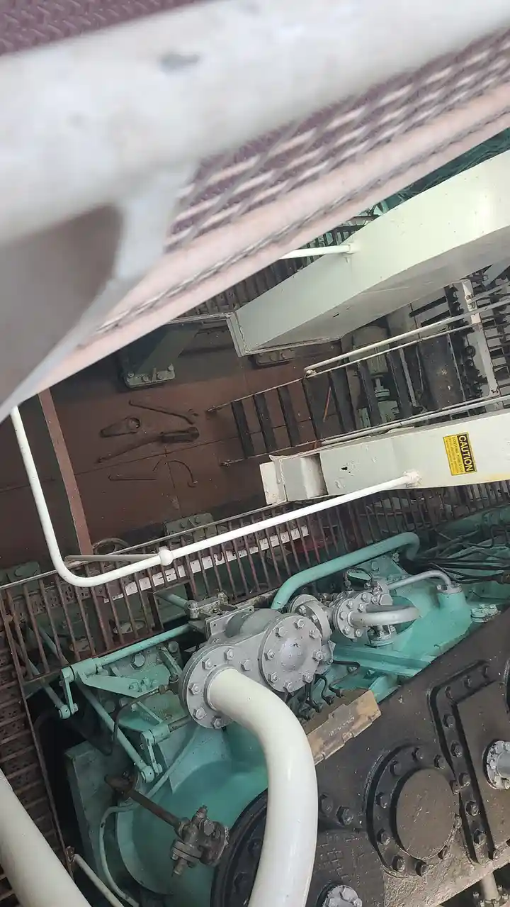 A low-angle shot of a rusty industrial engine room with teal-painted machinery, white pipes, and metal grates. Tools hang on…