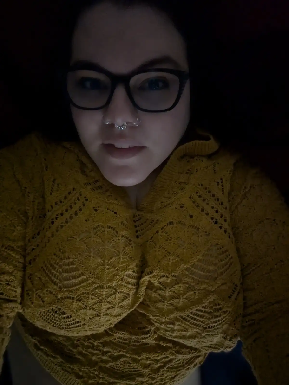 A person with dark hair, black-rimmed glasses, and a silver nose ring looks directly at the camera in a dimly lit room…