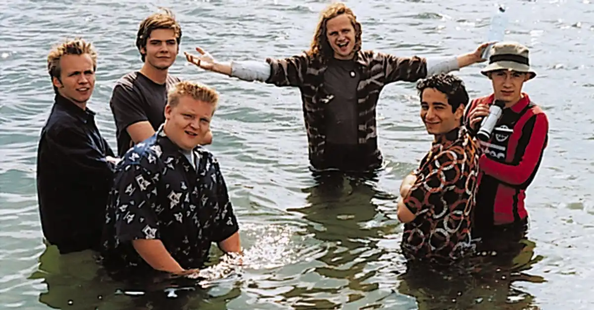 five young men stand waist-deep in murky water, smiling or posing. One man in the center with long hair and a plaid shirt…