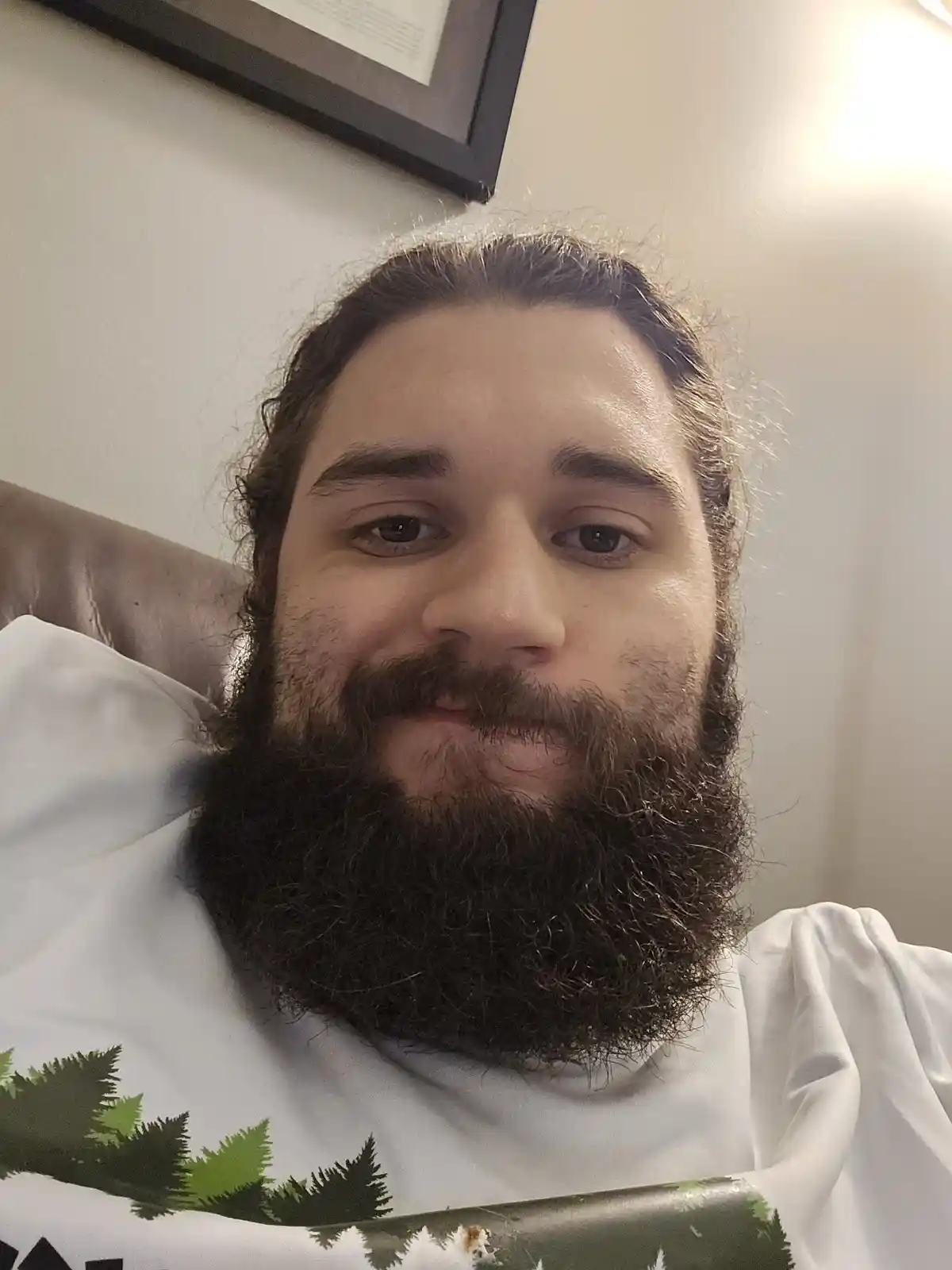 A man with a full, dark beard and long hair looks at the camera with a slight smirk while wearing a white t-shirt with a…