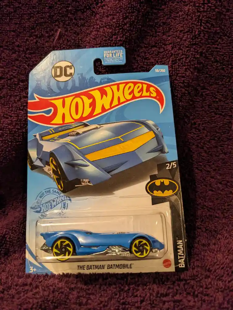 A Hot Wheels toy car package for 'The Batman' Batmobile, featuring a blue and yellow vehicle, is shown on a purple carpet…