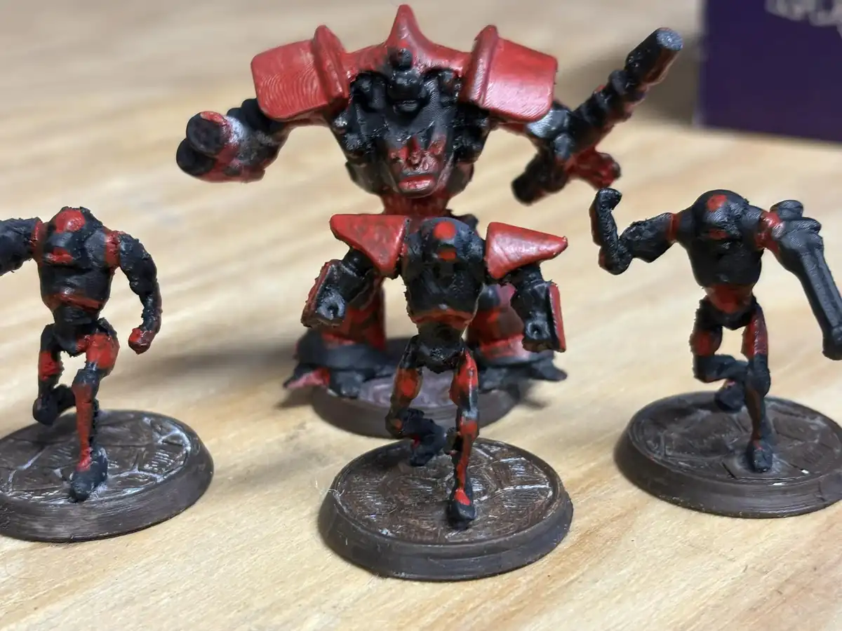 Three 3D-printed fantasy miniatures, painted black and red, stand on circular bases on a wooden surface. The central figure…