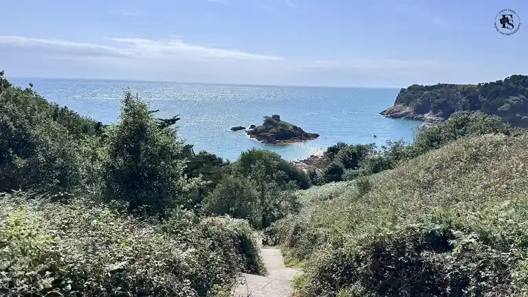 A scenic coastal view shows a winding stone path descending through lush green foliage toward a cove with turquoise water, a…