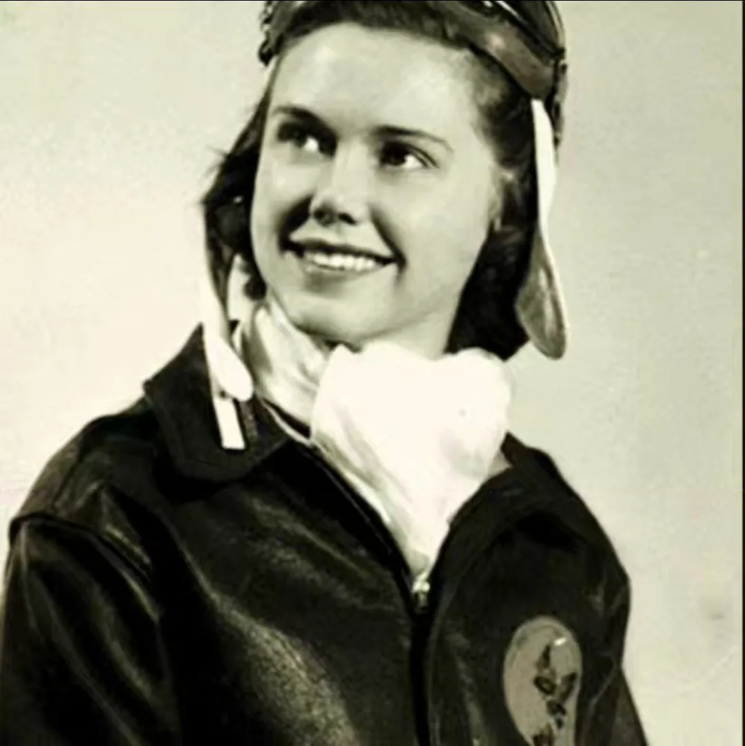 A vintage black-and-white photo shows a smiling young woman wearing a leather flight jacket, a white scarf, and a leather…