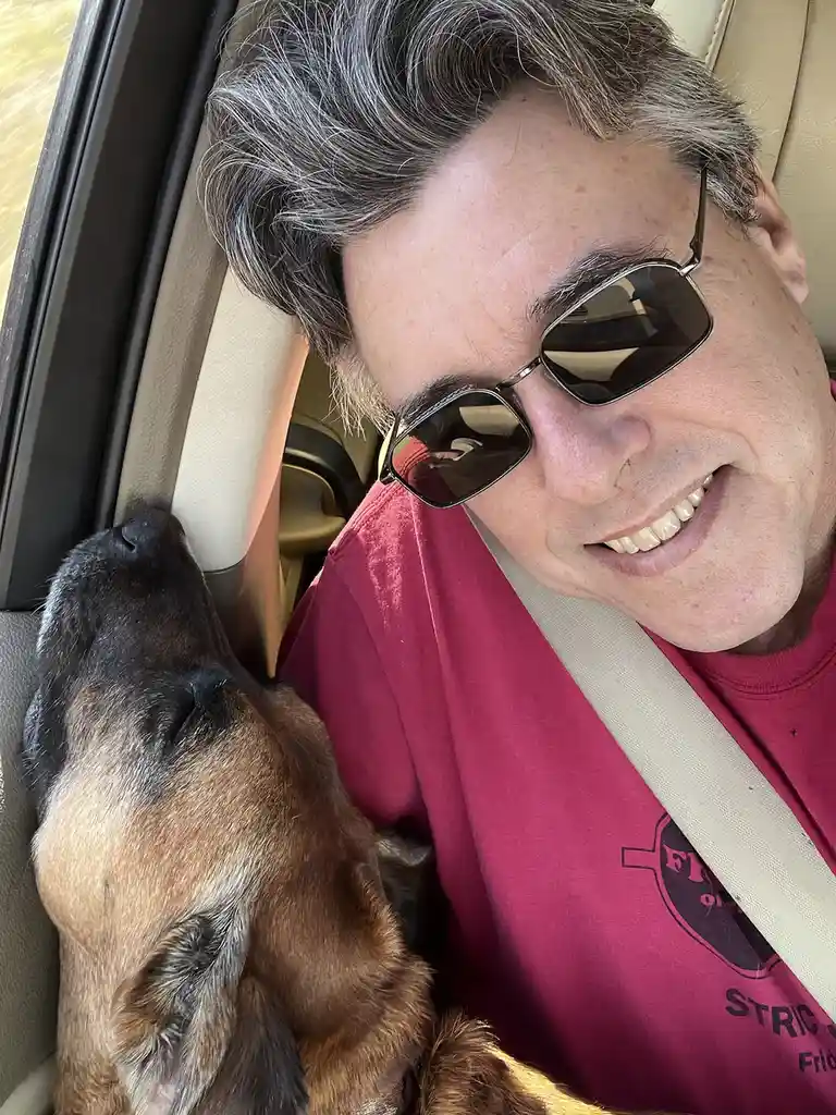 A man with gray hair and sunglasses smiles while wearing a seatbelt in a car, next to a brown and black dog resting its head…