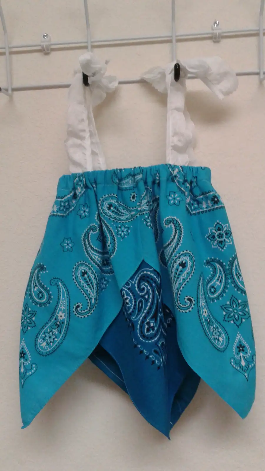 A turquoise bandana with black and white paisley patterns is fashioned into a dress, hanging on a white wire rack with white…