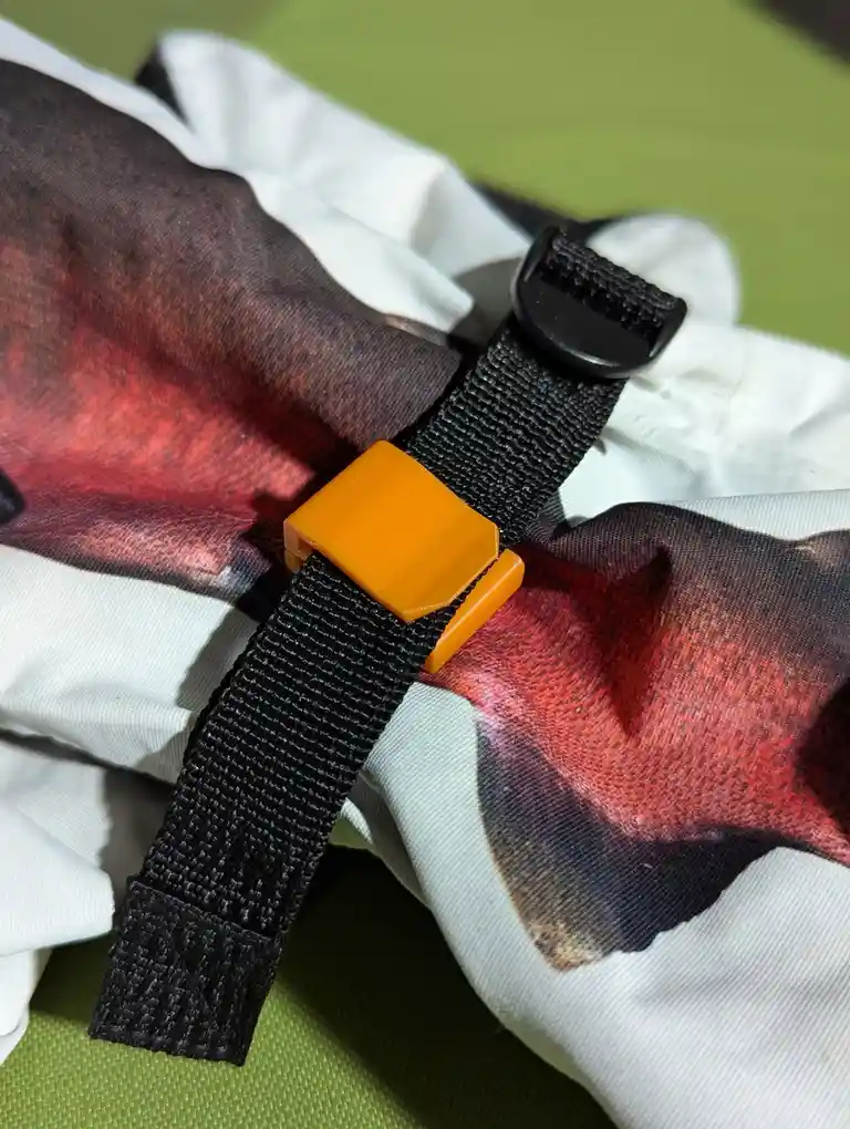 A close-up photo shows a black webbing strap with a bright orange plastic buckle fastened over a white fabric item printed…