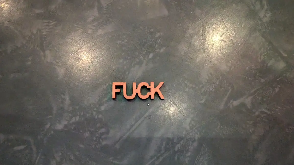The image shows the word 'FUCK' spelled out in orange, three-dimensional letters resting on a dark, textured…