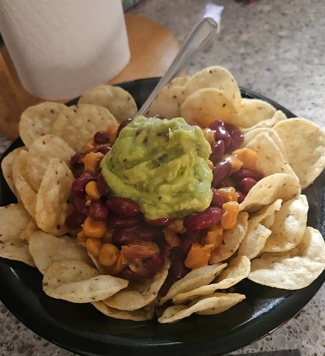A black bowl filled with tortilla chips, red kidney beans, corn, and a large dollop of guacamole in the center, with a spoon…