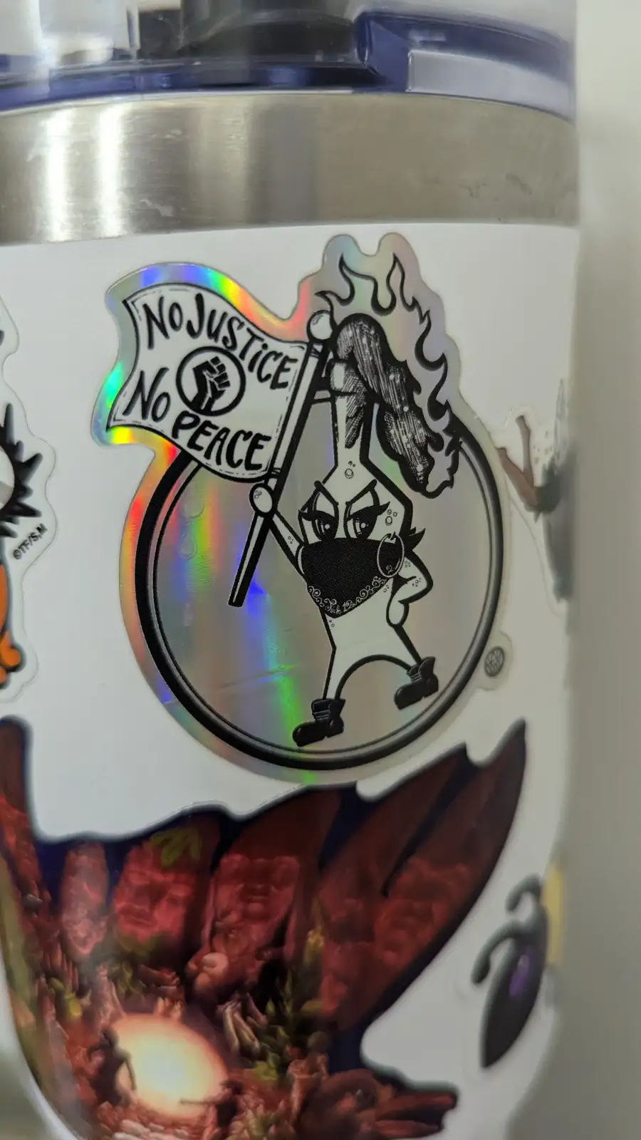 A close-up of a white tumbler with multiple stickers, prominently featuring a holographic sticker of a cartoon character…
