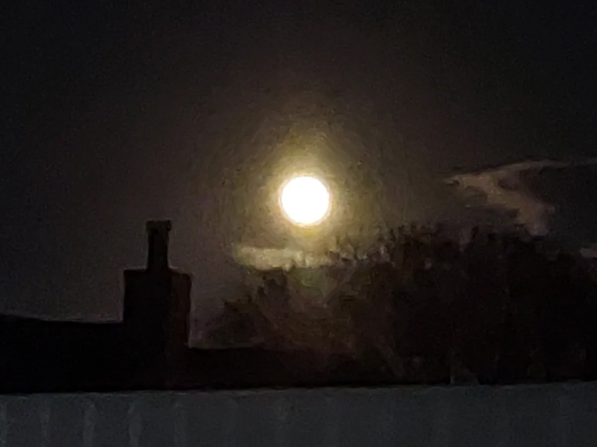 A blurry, low-resolution nighttime photo shows a bright, full moon glowing through a hazy or foggy sky, with dark…