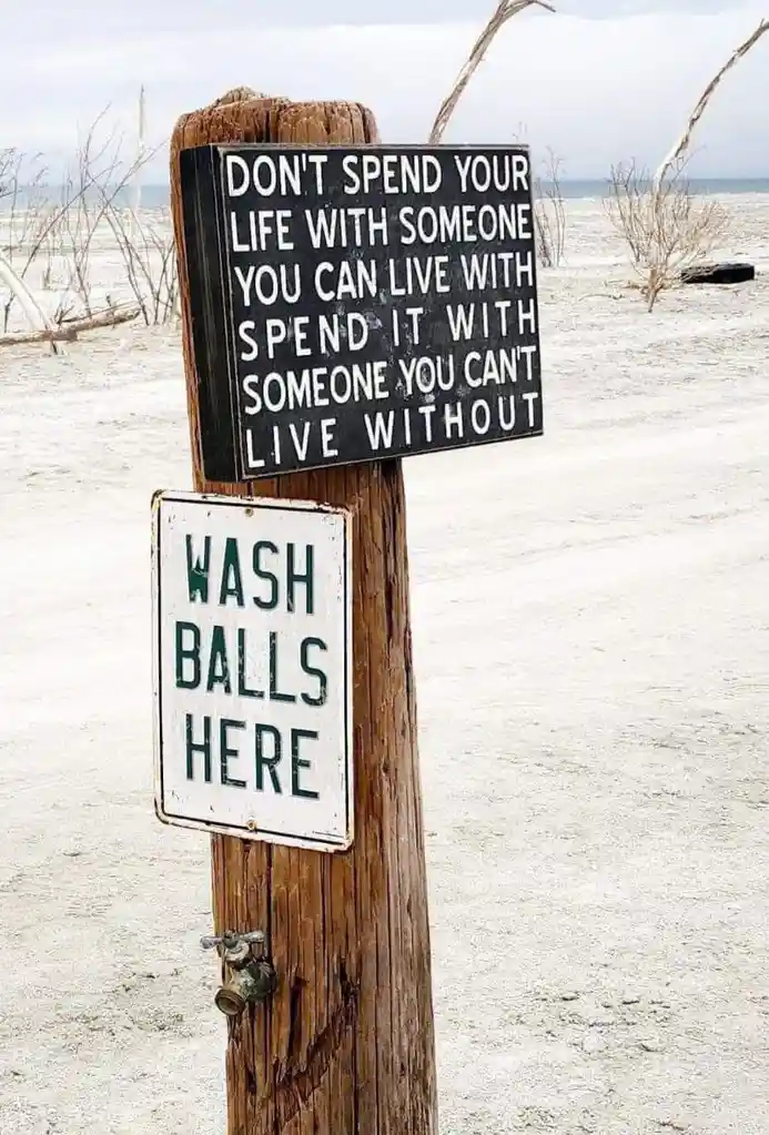 A weathered wooden post on a sandy beach holds two signs: the top black sign reads 'DON'T SPEND YOUR LIFE WITH SOMEONE YOU…