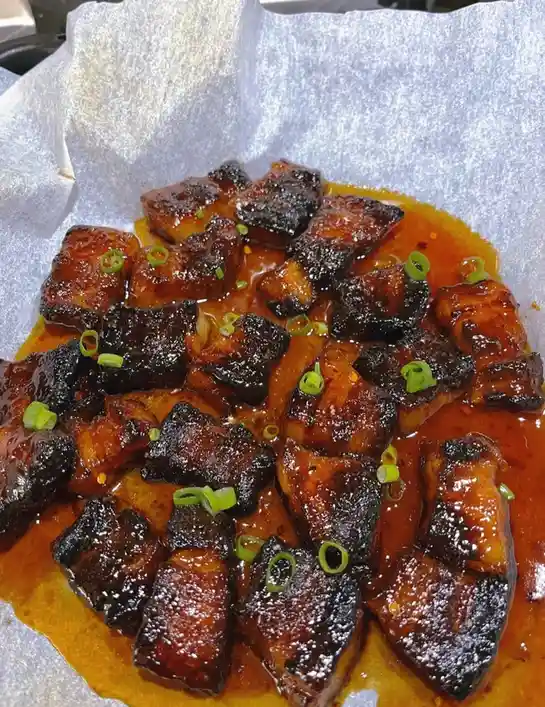 a close-up photo of glossy, dark-brown braised pork belly pieces in a rich sauce, garnished with chopped green onions…