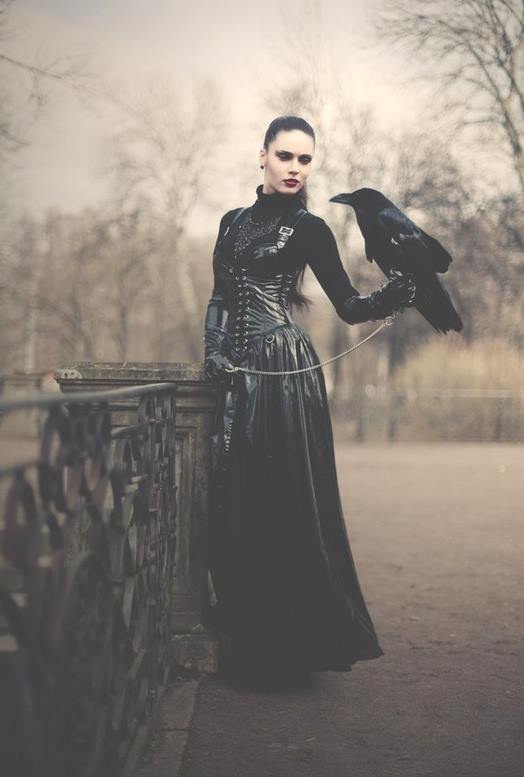 A woman in a black gothic dress with a corset and gloves stands outdoors holding a black crow on a leash. She has dark…