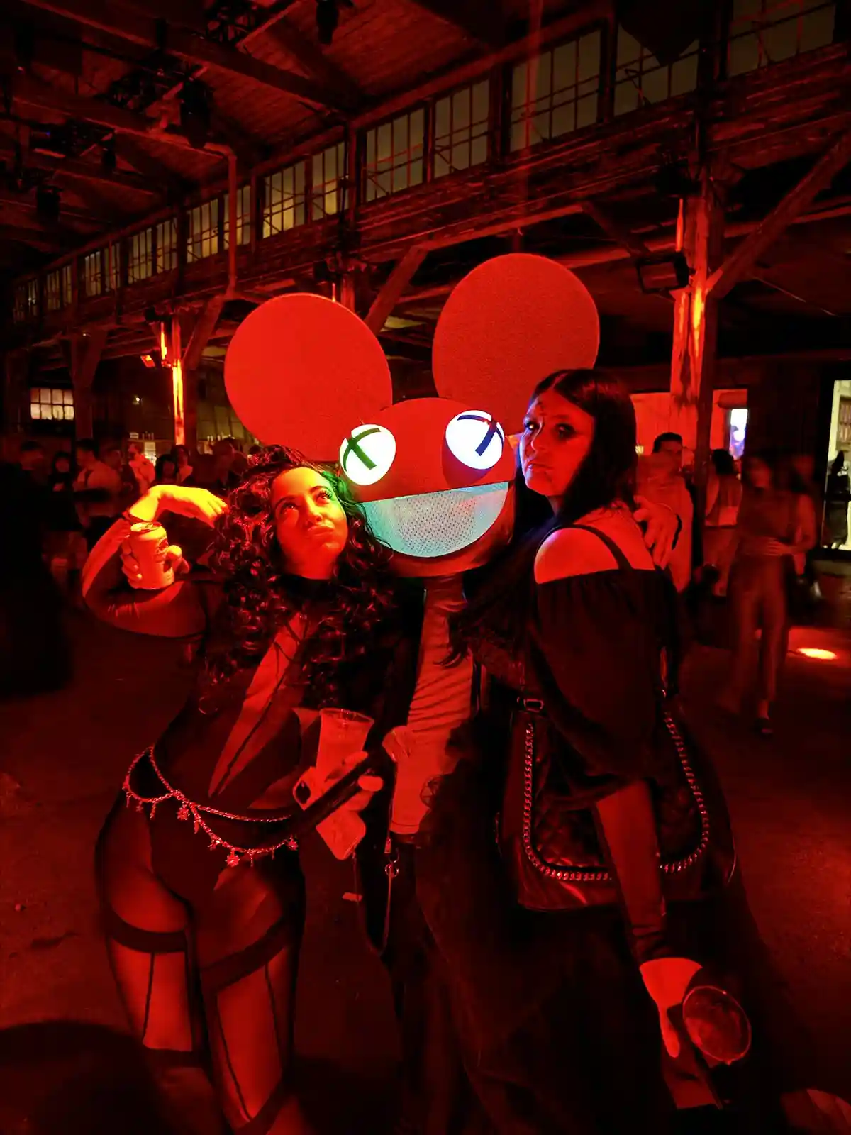 Three people pose together at a red-lit event; one wears a large, glowing red mouse head costume with white eyes marked with…