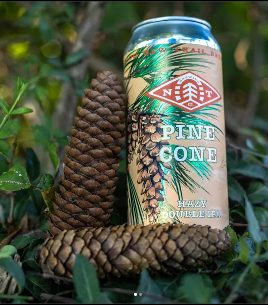 A beer can labeled 'Pine Cone Hazy Double IPA' from 'New Trail Brewing' is placed outdoors among real pinecones and green…