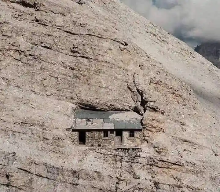 a small, weathered stone house is visibly embedded into the side of a steep, rocky mountain, with a dark roof and small…