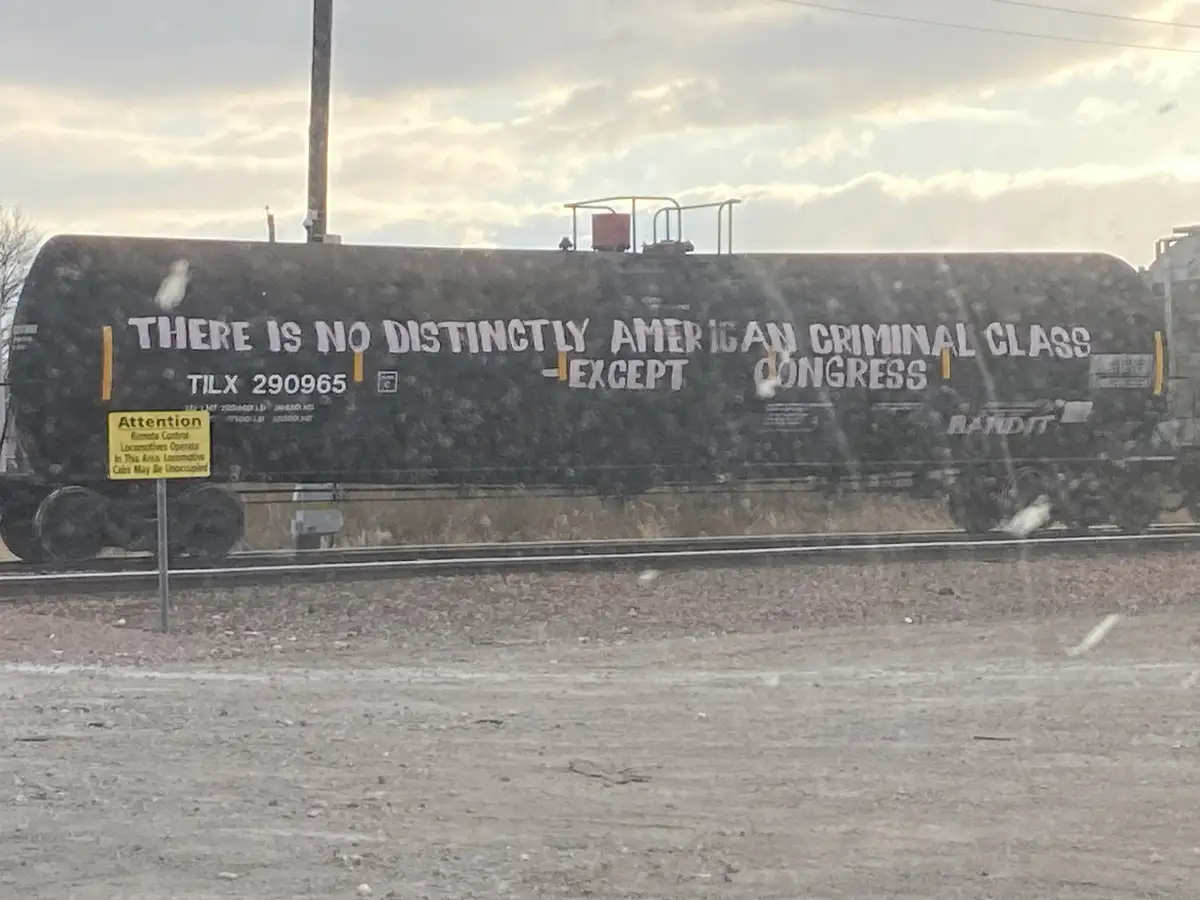 A black tanker railcar with white graffiti text reading 'THERE IS NO DISTINCTLY AMERICAN CRIMINAL CLASS -EXCEPT CONGRESS'…