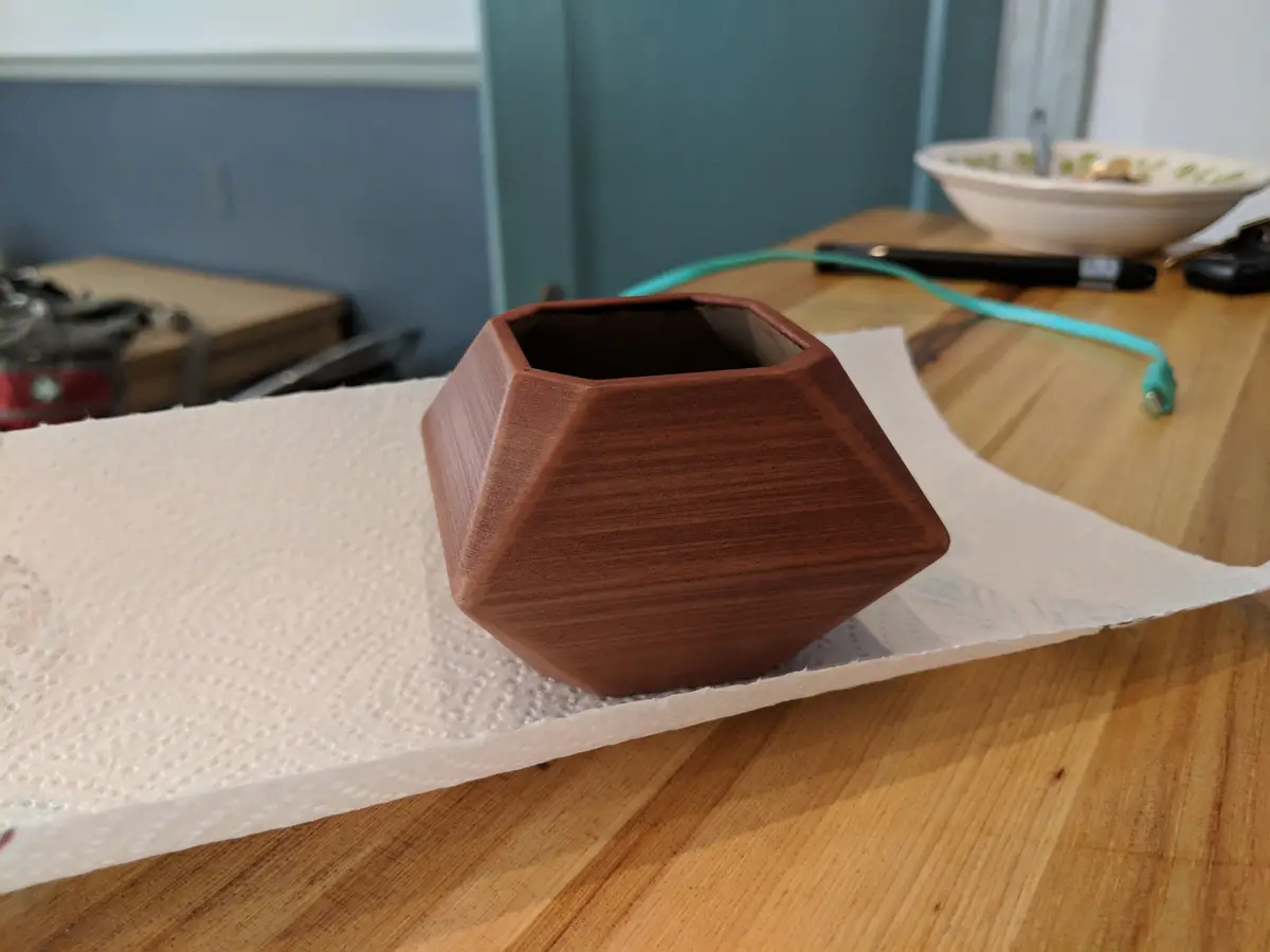 A brown, geometrically shaped 3d-printed vase sits on a white paper towel on a wooden table. The vase has visible layer…